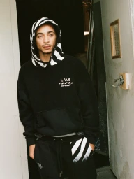 Model Julez Smith wearing a black Off-White hoodie with a striped hood and matching sweatpants standing in an open doorway.