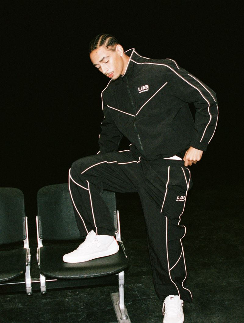 Model wearing a black piped tracksuit and white sneakers posing with one foot on a black chair against a dark background.