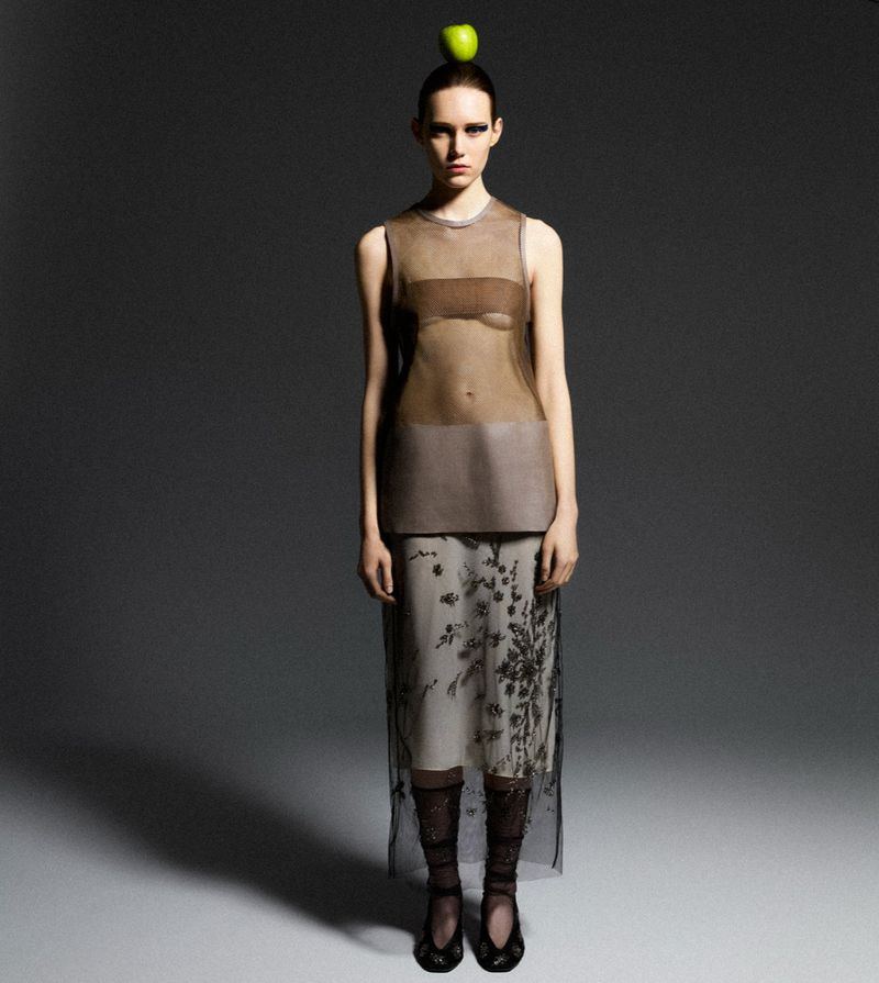 A slender fashion model poses in a sheer top and skirt, balancing a green apple on her head, against a dark grey studio backdrop.