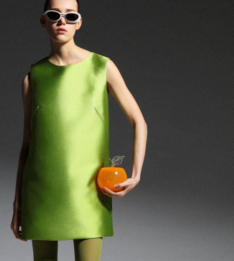 Fashion shot of a posing model wearing a lime-green mini dress, matching tights, white sunglasses, and holding an orange apple purse.