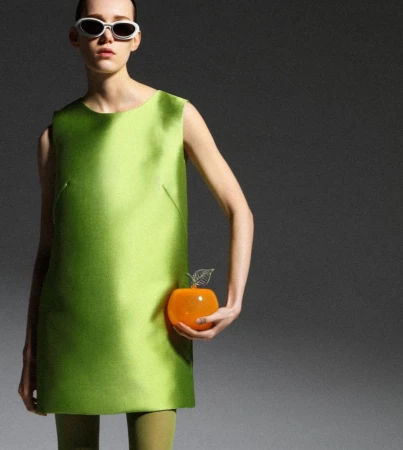 Fashion shot of a posing model wearing a lime-green mini dress, matching tights, white sunglasses, and holding an orange apple purse.