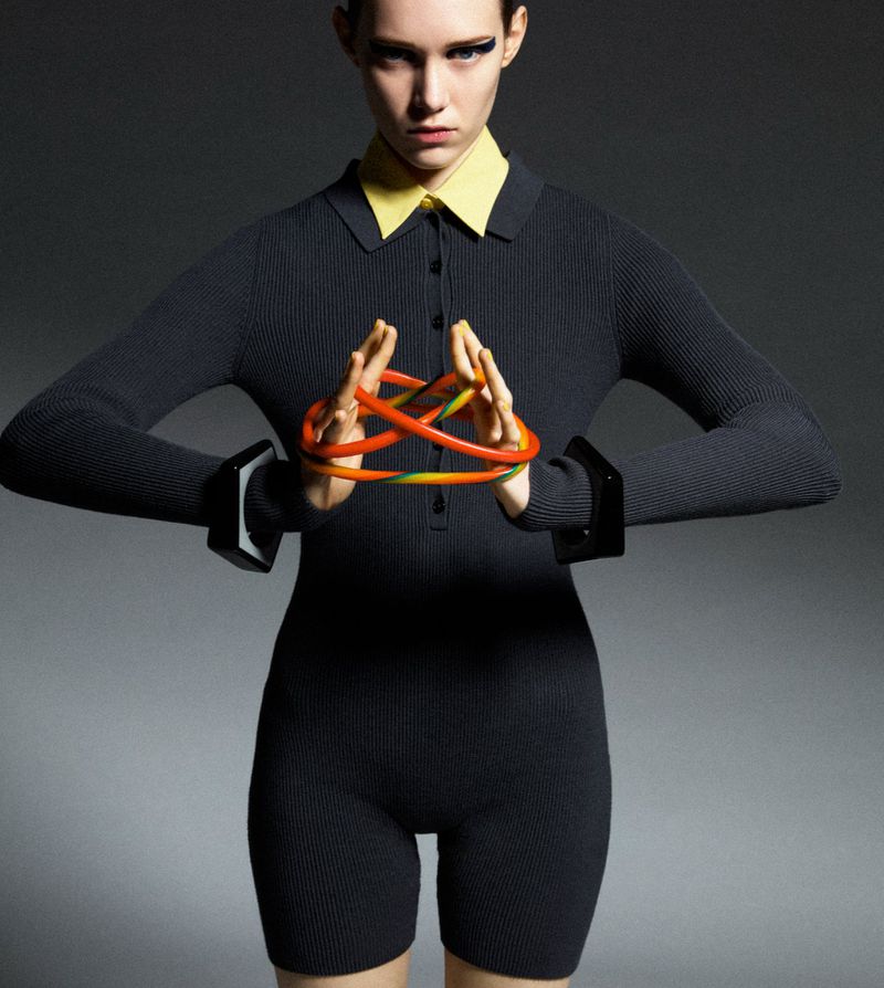 Fashion shot of a slim woman in a dark one-piece ribbed garment with a yellow collar and black wrist cuffs holding a circular object.