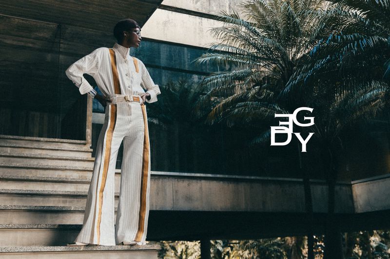 Fashion model in a cream wide-leg jumpsuit with tan stripes standing on concrete stairs beside palm trees.