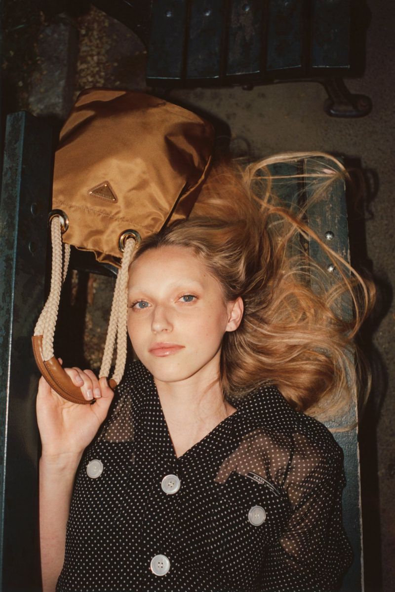 A young woman with blonde hair and a polka dot blouse poses with a brown Prada handbag against a dark background.
