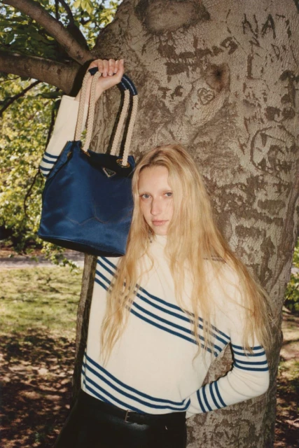 A woman in a striped sweater poses with a blue Prada handbag in front of a large tree in an outdoor setting with natural light.