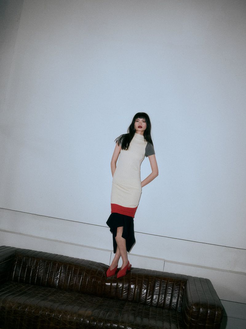 Chun Jin wearing a cream, red, and black color-blocked dress and red heels while standing on a dark leather sofa against a white wall.