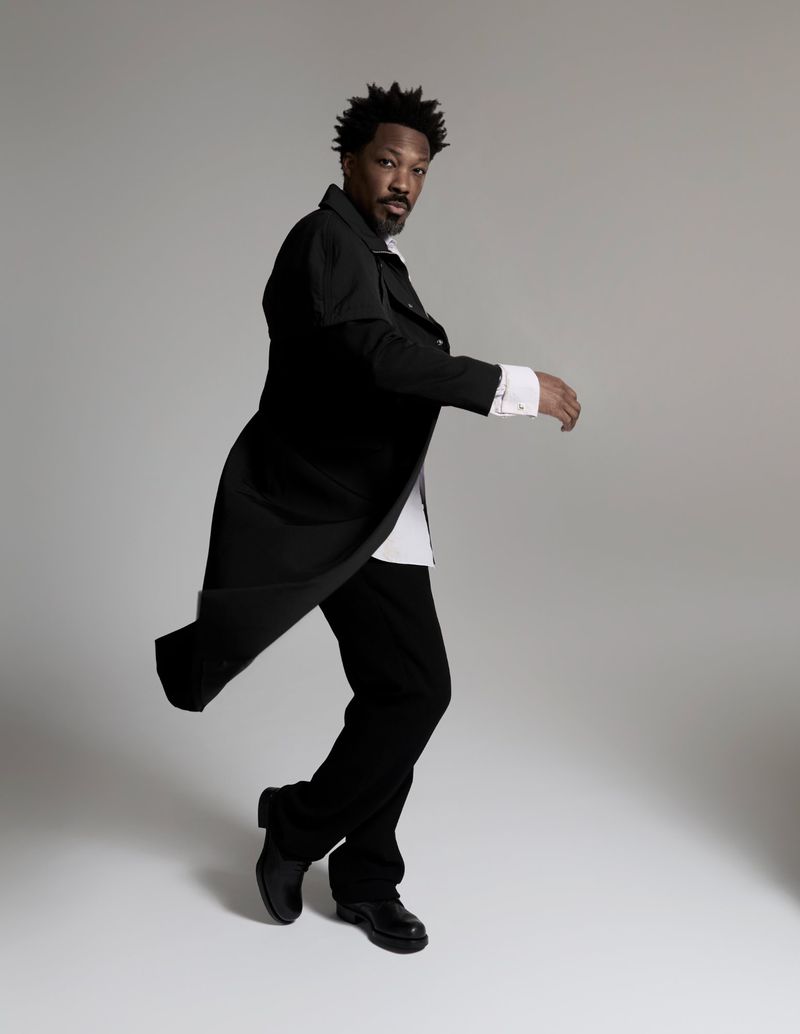 Corey Hawkins poses mid-motion wearing a flowing long black coat, white shirt, and black trousers against a neutral grey studio backdrop.