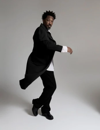 Corey Hawkins poses mid-motion wearing a flowing long black coat, white shirt, and black trousers against a neutral grey studio backdrop.