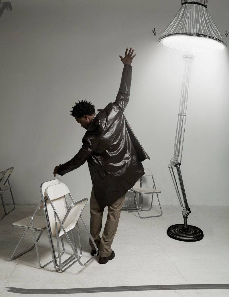 Actor Corey Hawkins posing in a dark glossy jacket with one arm raised toward an illustrated floor lamp among folding chairs.