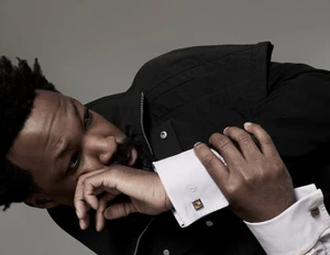 Corey Hawkins wearing a black jacket and white French cuff shirt with square cufflinks against a studio backdrop.