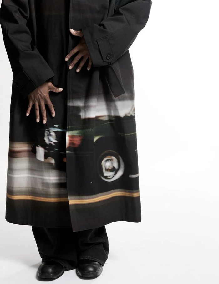 Cropped studio portrait of actor Corey Hawkins wearing a long black coat with a car graphic print, black trousers, and black leather shoes.