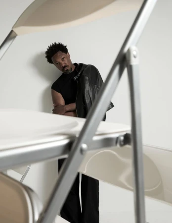 Actor Corey Hawkins in a black sleeveless shirt and leather jacket framed through folding chairs in a studio.