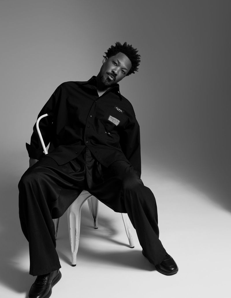 Actor Corey Hawkins seated on a white chair wearing an oversized black shirt and trousers in a black and white editorial portrait.