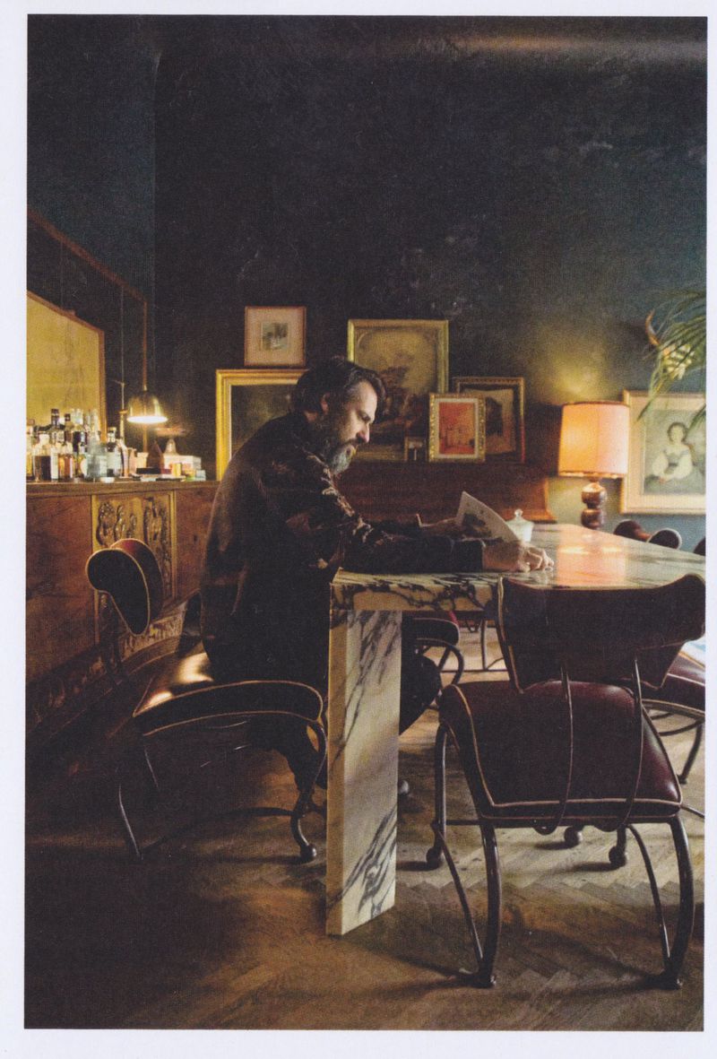 Man with a beard sitting at a marble table reading papers in a moody, art-filled room with dark walls.