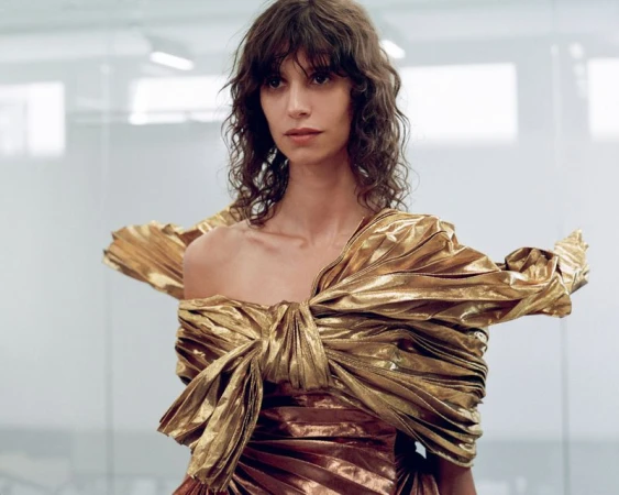 Model with dark wavy bangs wearing a sculpted metallic gold draped off-the-shoulder gown against a neutral studio backdrop.