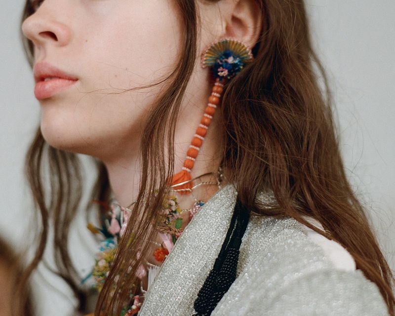Close-up profile of a model wearing a long beaded tassel earring, floral necklaces, and a shimmering silver garment.