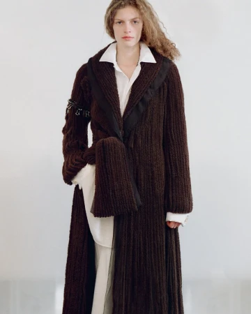 Model wearing a long textured dark brown knit coat over a white collared shirt against a white backdrop.