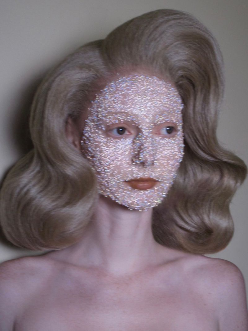 Model with sculpted blonde waves and a face covered in iridescent micro-beads against a plain neutral background.