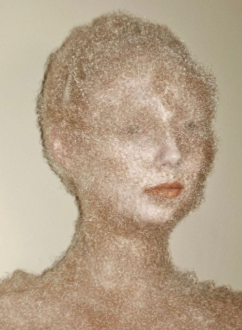 Close-up portrait of a model completely covered in fine fuzzy hair fibers with glossy nude lips against a neutral background.