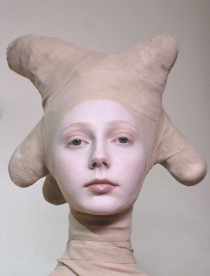 Close-up portrait of a model with pale makeup wearing a sculptural nude bandage headpiece and neck wrap against a neutral backdrop.
