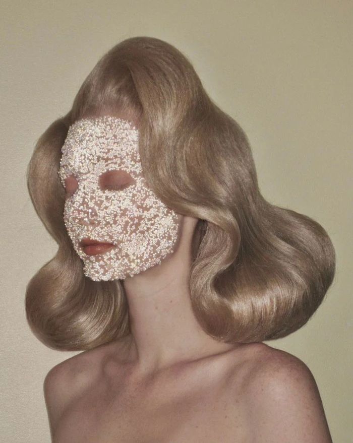 Model with voluminous sculpted retro blonde hair and a beaded pearl face covering against a neutral studio backdrop.