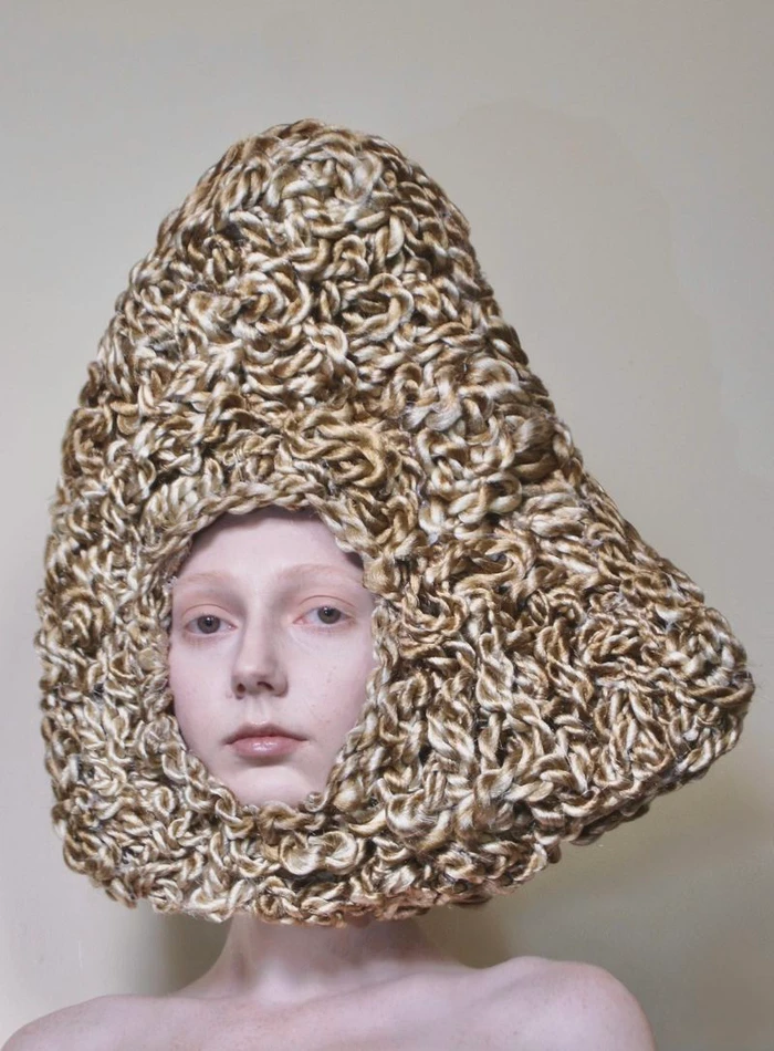 Close-up portrait of a woman with bare shoulders wearing a large triangular sculptural headpiece made of textured blonde and brown hair.