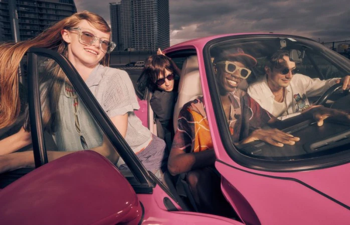 Four young adults ride in a pink sports car; an outdoor shot showcases urban architecture in the background, fashion and youthful energy.