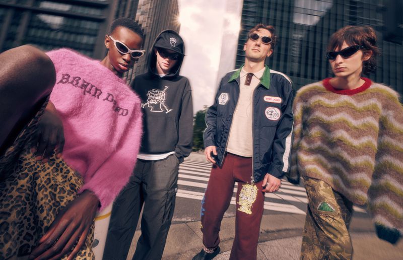 Four fashionably dressed adults stand outside a building wearing sunglasses, sweaters, a jacket, pants, and sneakers on a crosswalk.