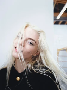 Close-up portrait of Anya Taylor-Joy with platinum blonde hair blown across her face wearing a black jacket with a gold button.