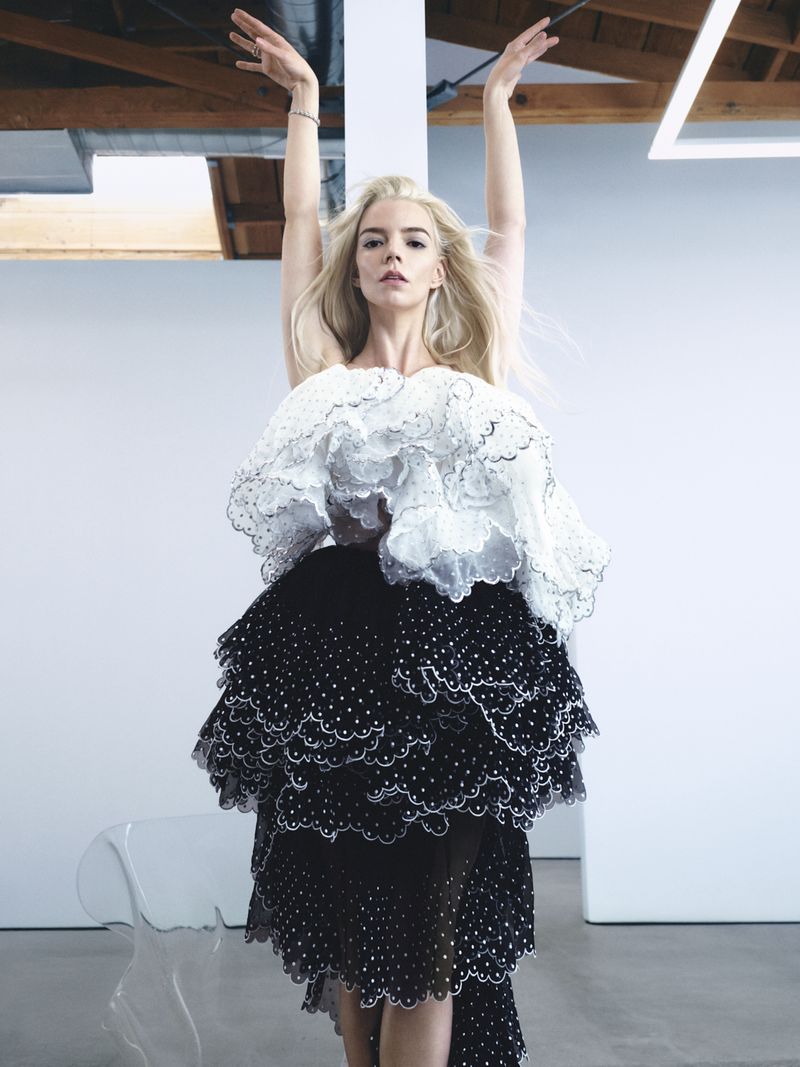 Anya Taylor-Joy poses with arms raised above her head, wearing a tiered black and white ruffle dress in a bright studio setting.