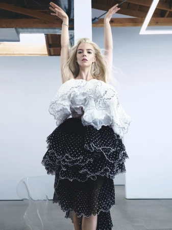 Anya Taylor-Joy poses with arms raised above her head, wearing a tiered black and white ruffle dress in a bright studio setting.