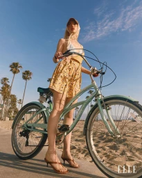 Chloe Cherry posing with a mint green cruiser bicycle on a sunny seaside path with palm trees in the background.