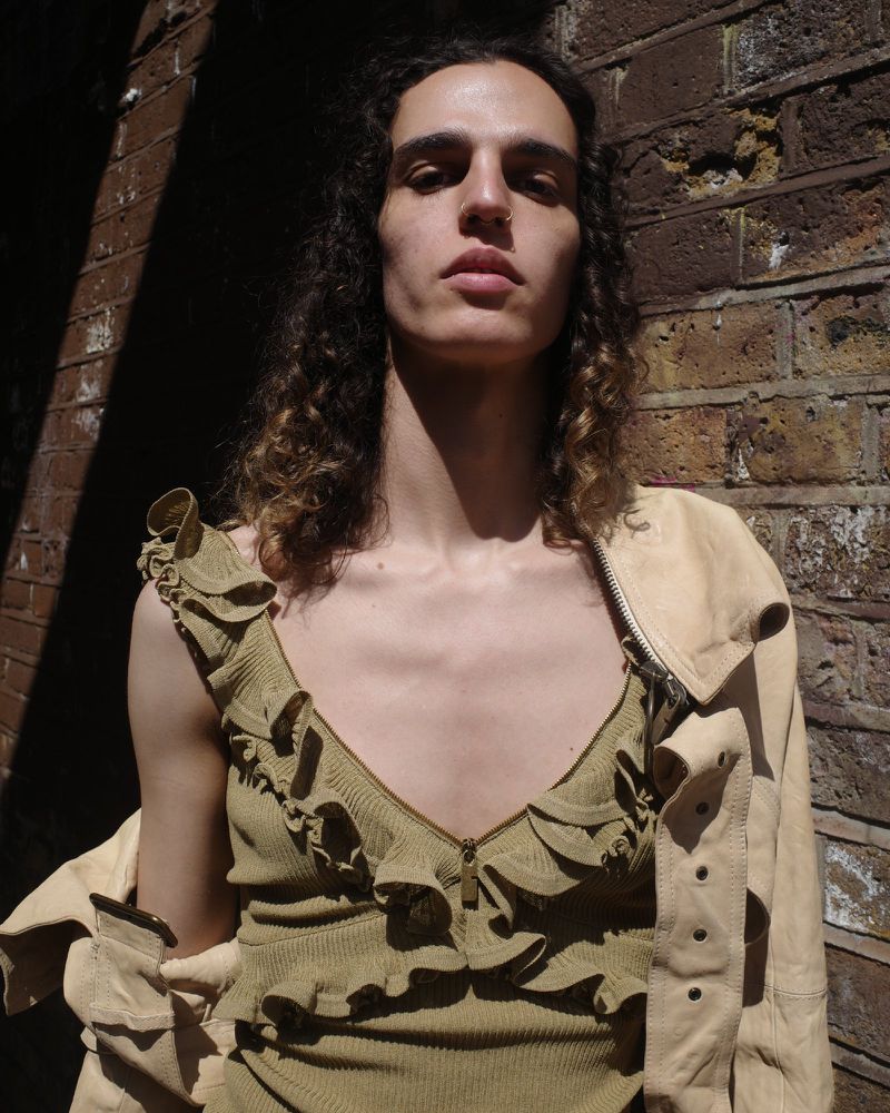 Close-up portrait of a young adult with long wavy hair, wearing a ruffle-trimmed top and jacket, standing against a brick wall.