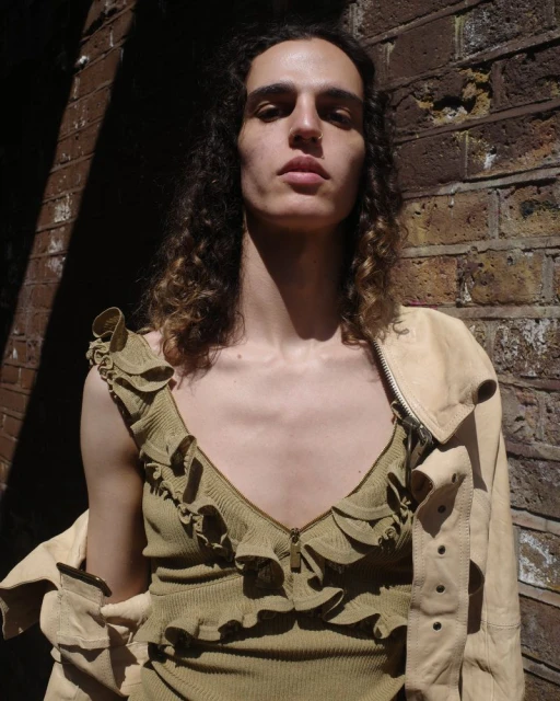 Close-up portrait of a young adult with long wavy hair, wearing a ruffle-trimmed top and jacket, standing against a brick wall.