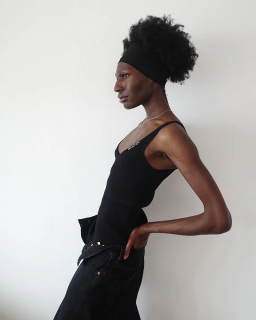 Black adult stands with hand in pants pocket in an eye-level shot in front of white wall in fashion pose, studio lighting.