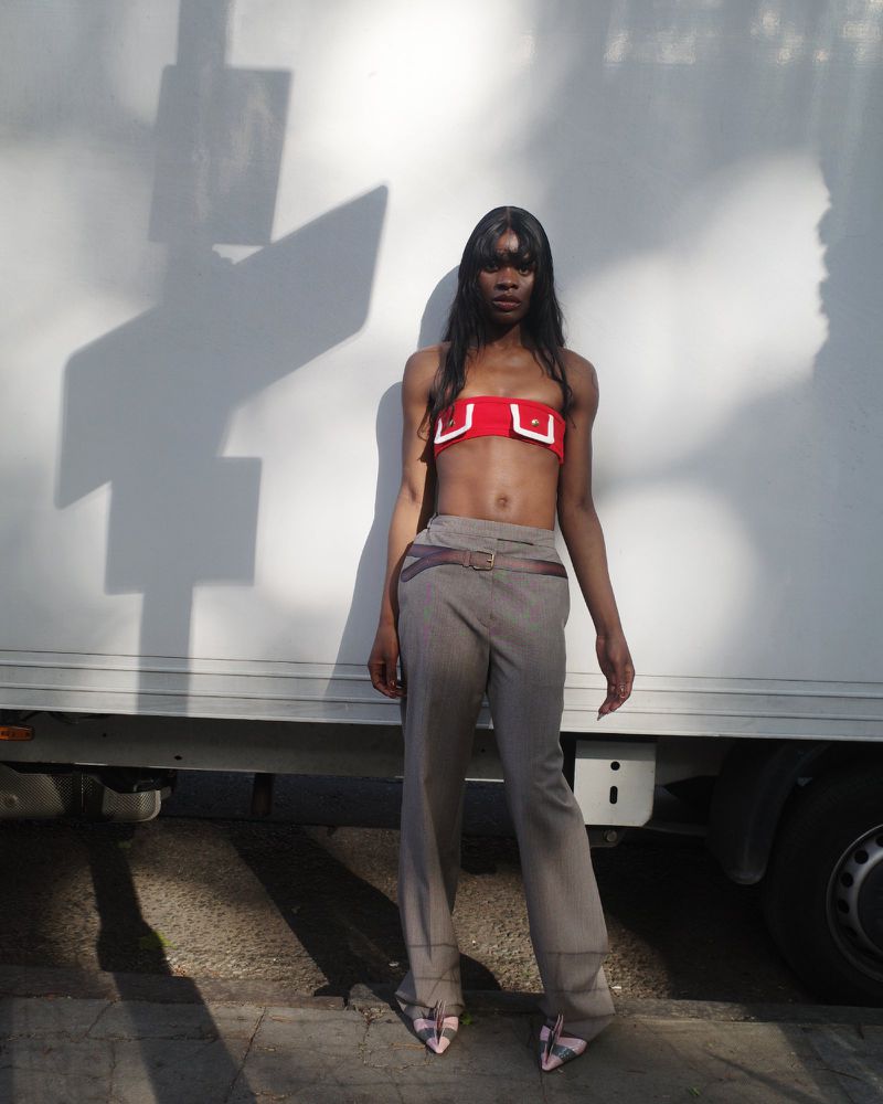 A slender Black woman stands outdoors in a red strapless top and taupe pants with a brown belt, posing with hands at her sides.