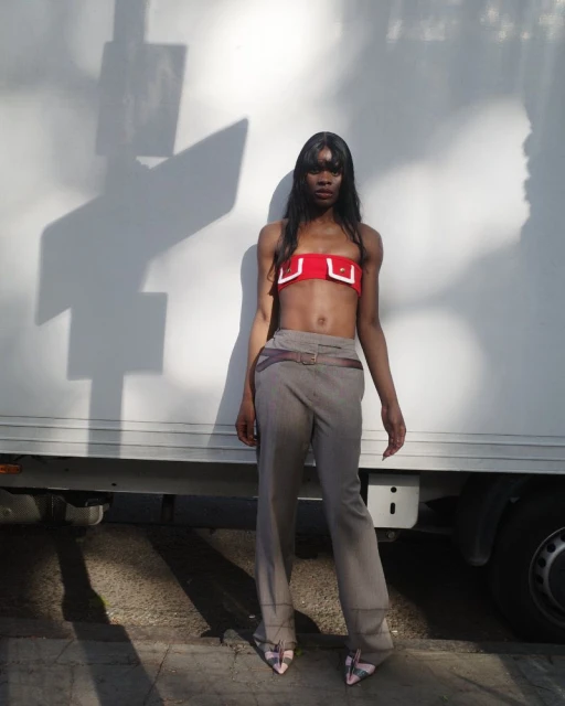 A slender Black woman stands outdoors in a red strapless top and taupe pants with a brown belt, posing with hands at her sides.