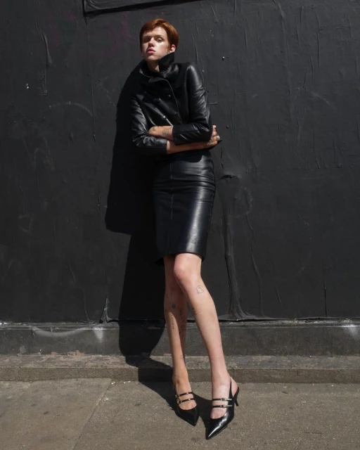 A redhead woman in a black leather jacket and pencil skirt poses against a dark wall, accessorized with heeled shoes.