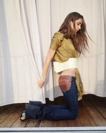 A young woman poses in an olive top, jeans, and a wide white belt in front of a draped white curtain indoors on a wooden floor.