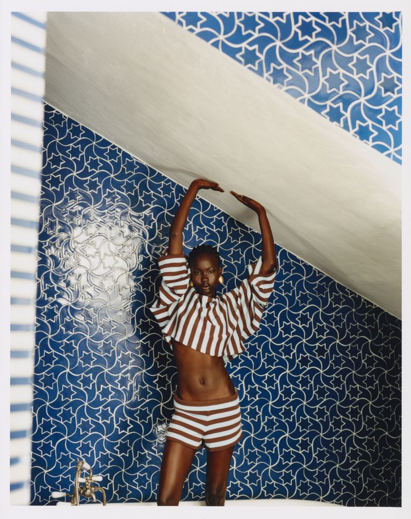 A woman posing with arms overhead in a bathroom with blue and white geometric star patterned tiled walls, wearing a brown striped set.