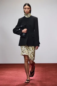 Fashion model walking on a red floor wearing a structured black jacket over a high-neck floral dress and carrying a dark clutch.