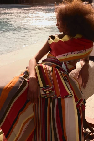 A woman in a colorful, striped dress poses near a beach, bathed in soft light, showcasing a blend of fashion and natural beauty.
