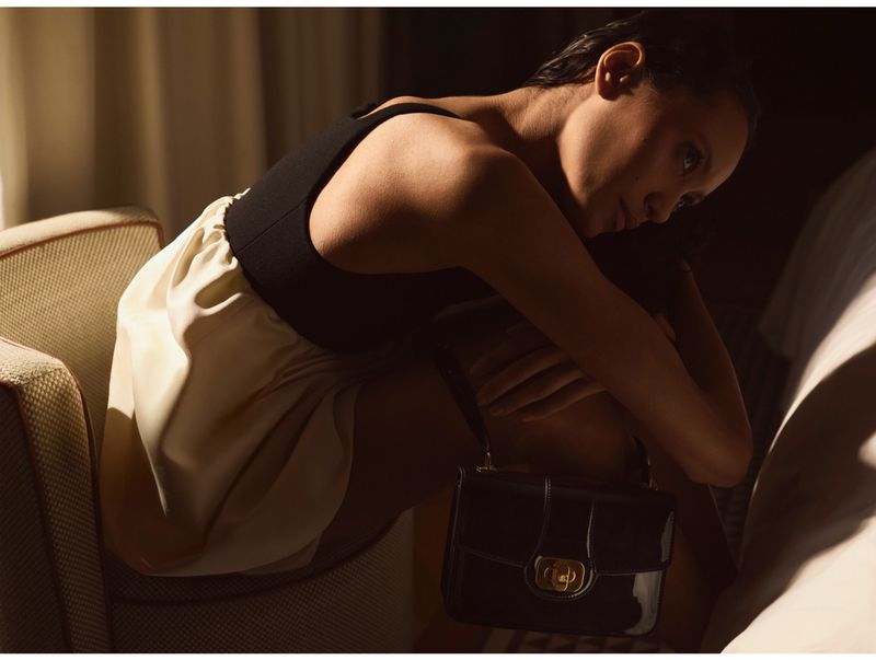 Adult female model in a black top and light skirt poses while sitting in an armchair next to a shiny black handbag indoors.
