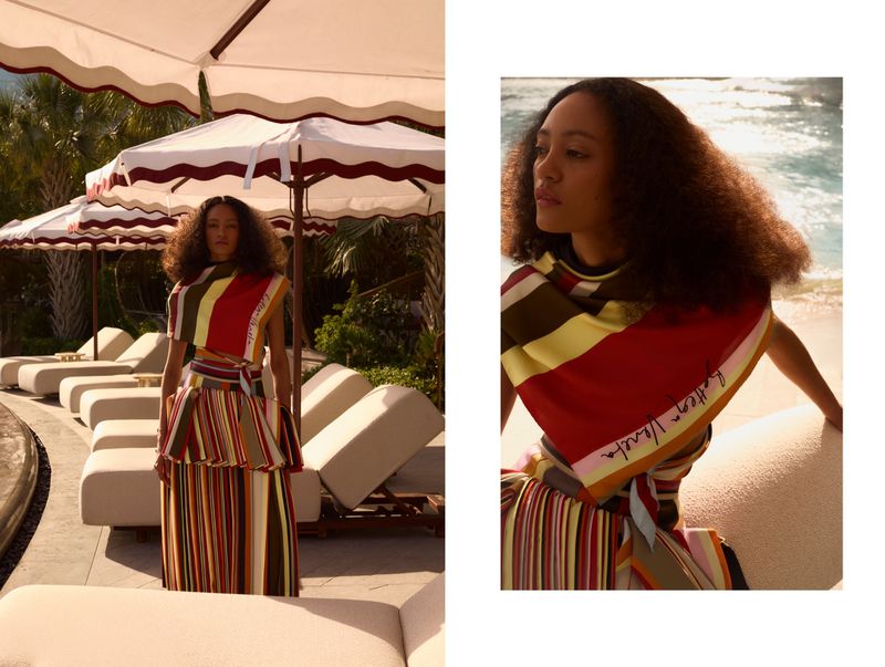 A young woman models a stylish striped dress and matching shawl next to an outdoor pool with lounge chairs under white umbrellas.