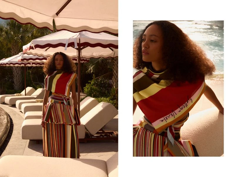 A young woman models a stylish striped dress and matching shawl next to an outdoor pool with lounge chairs under white umbrellas.