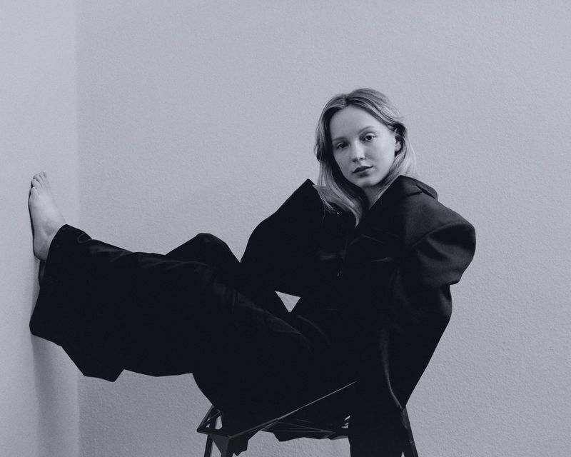 A young woman in a black oversized jacket and trousers reclines on a chair in front of a white wall in a minimalist portrait.