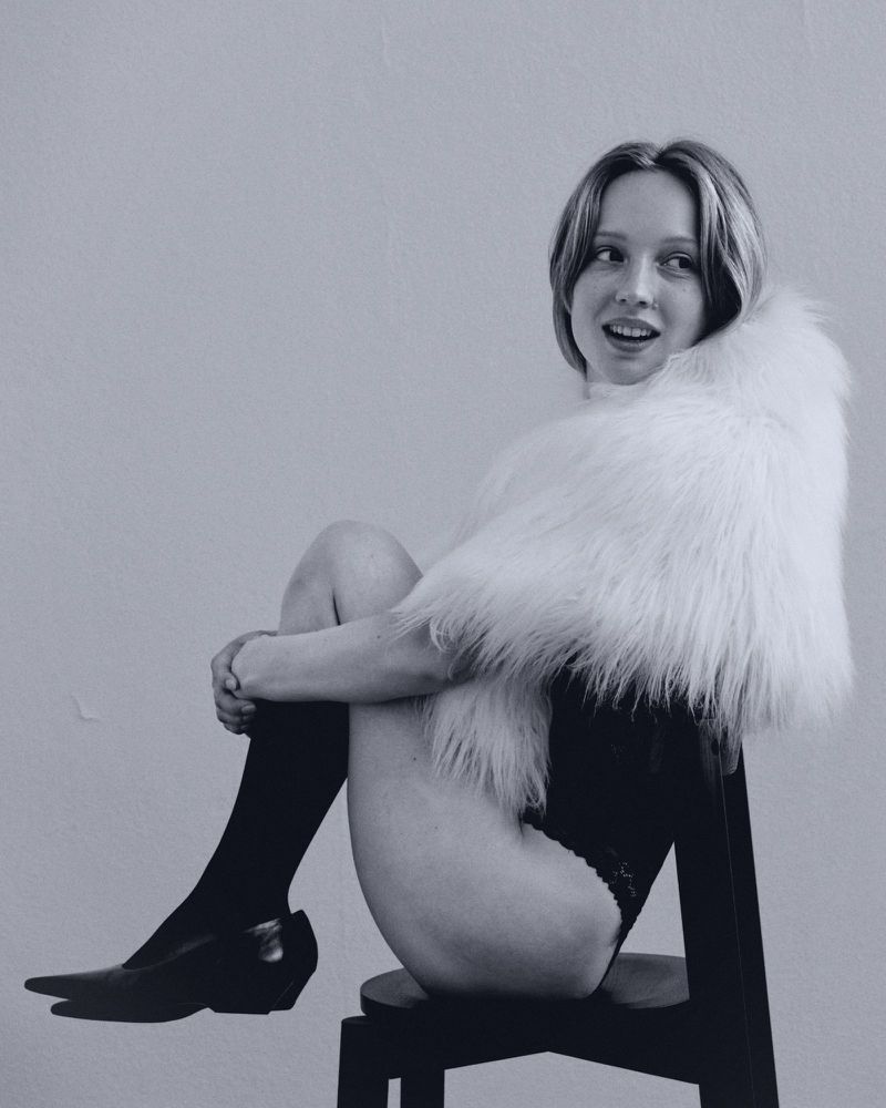 Monochrome portrait of a fair-skinned young woman with a short bob and a fur stole, sitting on a stool in front of a plain background.