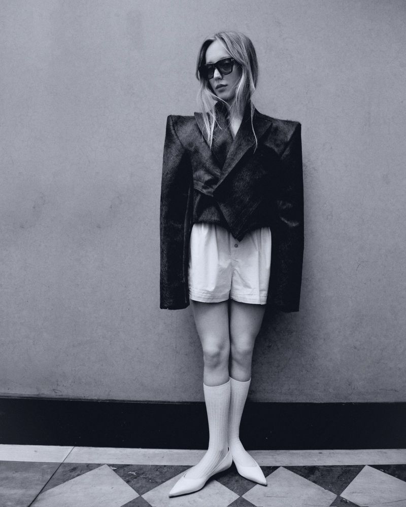 Black-and-white shot of a stylish woman in a blazer, shorts, socks, and pumps, posing against a textured wall with a tiled floor.