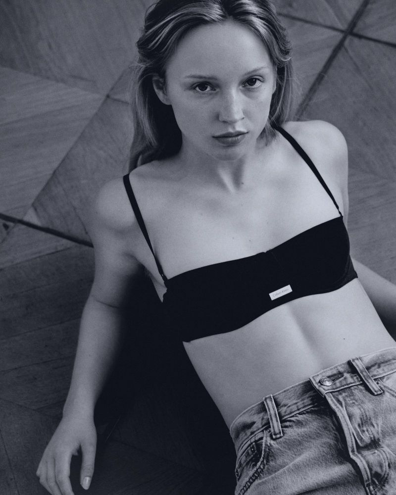 Portrait of a blonde woman in a black top and light blue jeans lying on a parquet floor in a studio with soft, diffused lighting.