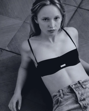 Portrait of a blonde woman in a black top and light blue jeans lying on a parquet floor in a studio with soft, diffused lighting.